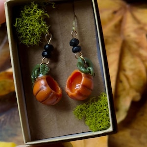 May include: A pair of pumpkin-shaped earrings with green stems and black beads hanging from gold hooks. The earrings are displayed in a small box with green moss.