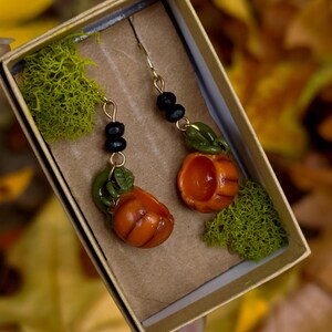 May include: A pair of pumpkin earrings with green leaves and black beads hanging from gold hooks. The earrings are displayed in a small box with green moss.