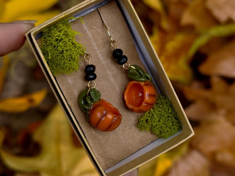 May include: A pair of pumpkin earrings with green leaves and black beads hanging from gold hooks. The earrings are displayed in a small box with green moss.