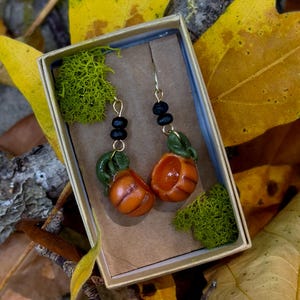 May include: A pair of gold pumpkin earrings with black beads and green leaves. The earrings are in a small box with green moss.