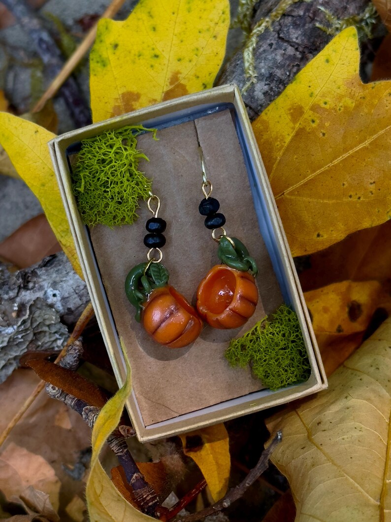 May include: A pair of gold pumpkin earrings with black beads and green leaves. The earrings are in a small box with green moss.