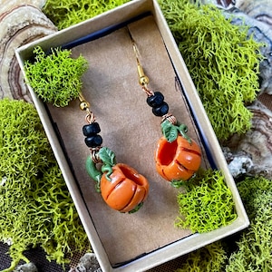 May include: A pair of pumpkin-shaped earrings with black beads and gold hooks. The earrings are displayed in a small cardboard box on a bed of green moss.