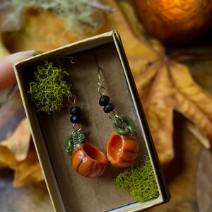 May include: A pair of pumpkin-shaped earrings with green stems and black beads hanging from gold-colored hooks. The earrings are displayed in a small box lined with green moss.