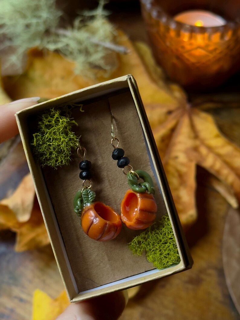 May include: A pair of pumpkin-shaped earrings with green stems and black beads hanging from gold-colored hooks. The earrings are displayed in a small box lined with green moss.