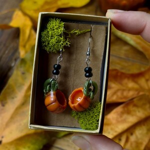 May include: A pair of pumpkin-shaped earrings with green stems and black beads hanging from silver hooks. The earrings are displayed in a small box with green moss.