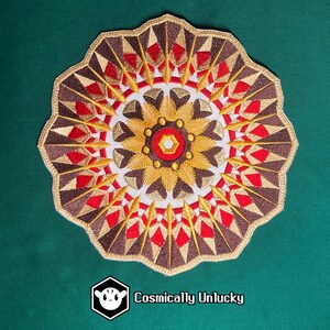 Peut inclure: Écusson brodé avec un motif mandala circulaire. Le patch présente un motif détaillé dans les tons marron, rouge, or et blanc. Le bord extérieur est souligné en or. Le texte "Cosmically Unlucky" est en bas.