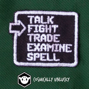 May include: Black embroidered patch with white text: "TALK FIGHT TRADE EXAMINE SPELL". The patch has a white border and an arrow design. The patch is on a green background. The "Cosmically Unlucky" logo is in the bottom left corner.