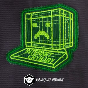 Peut inclure: Patch brodé vert représentant un ordinateur triste avec le texte "INTERNET CRYBABY" sur le clavier. Le patch est fabriqué par COSMICALLY UNLUCKY.