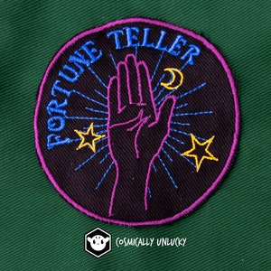 May include: A black and purple embroidered patch with the text "Fortune Teller" in blue. The patch features a stylised hand with a crescent moon and two stars.