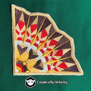 Peut inclure: Écusson brodé avec un motif géométrique rouge, marron, or et blanc. L'écusson a une bordure dorée et un motif stylisé en forme d'éventail. Le texte "Cosmically Unlucky" est imprimé en bas à droite.
