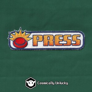 Peut inclure: Un patch brodé blanc et bleu avec le mot "PRESS" en lettres orange. Un bouton de dessin animé rouge et jaune se trouve sur le côté gauche du patch.