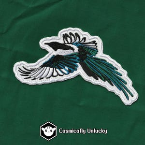 May include: A white, black and blue embroidered patch of a magpie in flight. The patch is on a green background.
