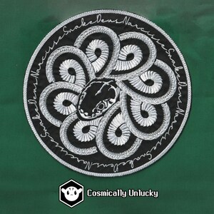 May include: A black and white embroidered patch with a circular design of snakes intertwined. The text "Snake Dens Narrow" is written around the edge of the patch.