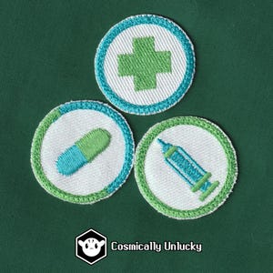 May include: Three white embroidered patches with green and blue accents. The patches feature a green cross, a blue and green pill, and a blue syringe.