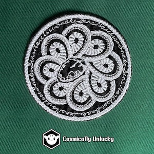 May include: Circular embroidered patch with a black background and white floral design. The patch features a central design with a snake-like figure and a border with white text. The patch is set against a green background.