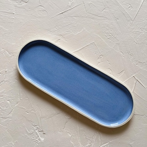 May include: A long, oval-shaped ceramic serving tray. The tray has a light blue interior and a white rim. The background is a textured, off-white surface. The tray is a decorative piece for serving food or displaying items.