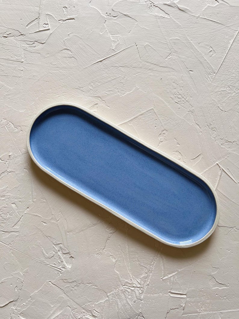 May include: A long, oval-shaped ceramic serving tray. The tray has a light blue interior and a white rim. The background is a textured, off-white surface. The tray is a decorative piece for serving food or displaying items.