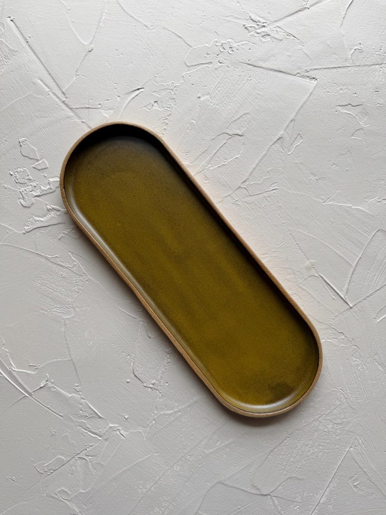 May include: An olive green ceramic serving tray with a rounded rectangular shape. The tray has a light brown rim and a smooth, glossy surface. The tray is approximately 12 inches long and 5 inches wide.