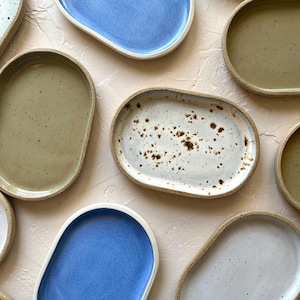 May include: Overhead shot of several handmade ceramic oval plates in various colors. The plates include shades of blue, white with brown speckles, and olive green, all with a natural, unglazed rim. The plates are arranged on a light pink textured surface.