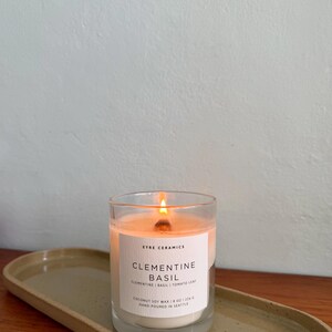 May include: A lit glass candle with a white label that reads "Clementine Basil" sits on a ceramic tray. The candle is made with coconut soy wax and hand-poured in Seattle. The candle is 8 oz.