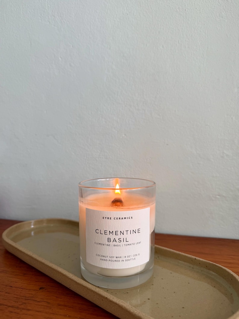 May include: A lit glass candle with a white label that reads "Clementine Basil" sits on a ceramic tray. The candle is made with coconut soy wax and hand-poured in Seattle. The candle is 8 oz.