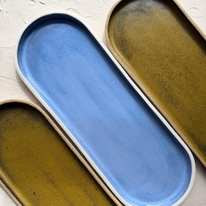 May include: Three elongated oval ceramic trays with raised edges. One tray features a smooth, matte light blue glaze, while the other two trays display a textured, mottled olive green finish. Each tray has a contrasting unglazed beige rim.