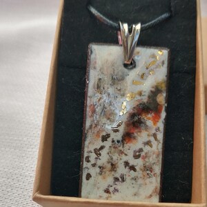 Peut inclure: Un collier pendentif rectangulaire avec un fermoir argenté et un cordon noir. Le pendentif présente un fond blanc avec des accents bruns, orange et dorés. Le collier est présenté dans une boîte marron avec un intérieur noir.