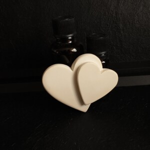 Peut inclure: Deux petits flacons en verre brun avec des bouchons noirs sont posés sur deux cœurs en céramique blanche. Les cœurs sont joints l'un à l'autre.