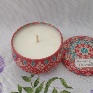 Peut inclure: Une boîte de bougie rouge avec une bougie blanche et une mèche à l'intérieur. La boîte a un motif de mandala coloré et les mots "Pomme Cannelle" écrits sur le couvercle.