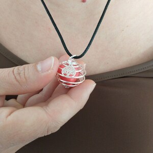 Könnte beinhalten: Eine Halskette aus silbernem Draht mit einem Käfig, in dem sich eine rote Kugel befindet. Der Käfig hat ein Lebensbaum-Design. Die Halskette ist an einem schwarzen Band.