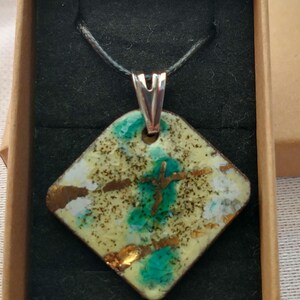 Peut inclure: Un collier pendentif carré avec un cordon noir. Le pendentif a un fond jaune clair avec des accents turquoise et dorés. Le pendentif est dans une petite boîte marron avec un intérieur noir.