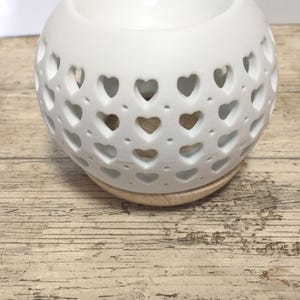 Könnte beinhalten: Ein weißer Keramik-Diffusor für ätherische Öle mit herzförmigen Aussparungen. Der Diffusor hat eine runde Form mit einer vertieften Oberseite für Öl und steht auf einem hellen Holzsockel. Das Design ist schlicht und elegant.