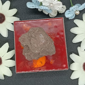 Peut inclure: Un carreau carré rouge brillant avec des accents orange, surmonté d'une roche volcanique brun foncé, rugueuse et poreuse. Le carreau est entouré de quatre fleurs en tissu blanc à centre brun foncé et de petites fleurs crochetées bleu clair.