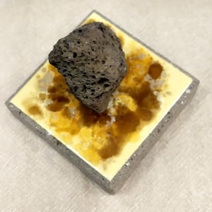 May include: A square, grey concrete base featuring a top surface with a yellow and brown mottled, resin-like finish. A single, dark grey, porous volcanic rock is positioned off-centre on the top of the square base, showcasing a rough texture.