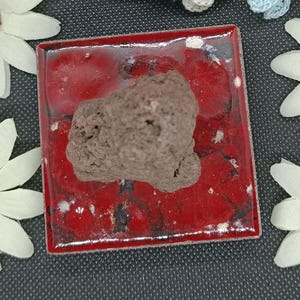 May include: A square, glossy red ceramic tile with a dark, porous, brown rock-like object resting in the centre. The tile features a crackled glaze finish with black accents visible beneath the vibrant red surface, creating a textured appearance.