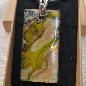 May include: A rectangular pendant with a silver bail, displaying an abstract design. The pendant combines olive green, cream, and hints of blue and gold. The pendant is presented in a small, open cardboard box.