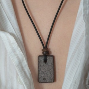 Op de afbeelding: Een zwarte leren koordsnoer met een rechthoekige hanger van bruine steen. De hanger heeft een ruw, getextureerd oppervlak.