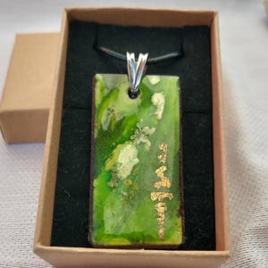 May include: A rectangular pendant with a green and gold design, presented in a brown gift box. The pendant has a silver-coloured bail and is suspended from a black cord. The green surface has gold leaf accents.