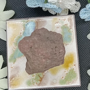 May include: A square ceramic tile with a glossy finish featuring abstract splashes of blue, yellow, and green glaze. A rough, porous, dark brown volcanic rock is centred on top of the tile. The tile has a dark brown edge and a flat surface.
