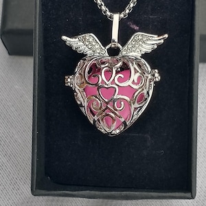 Può includere: Una collana con pendente a forma di cuore color argento con interno rosa, esposta in una scatola nera. Il pendente presenta un design traforato con volute decorative ed è ornato da piccole ali scintillanti.