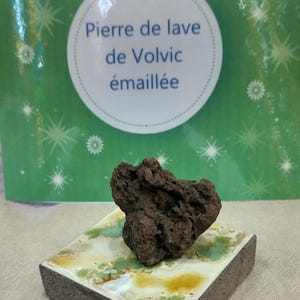 Op de afbeelding: Een donkerbruine vulkanische rots op een vierkante tegel met een witte, groene en gele glazuur. De tegel ligt op een beige oppervlak. De tekst op de groene achtergrond achter de rots luidt "Pierre de lave de Volvic émaillée".