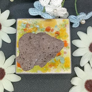 Peut inclure: Un carreau carré à la finition brillante présentant un motif tacheté jaune, orange et bleu clair. Une roche volcanique brun foncé, poreuse et irrégulière est centrée sur le carreau. Le carreau est entouré de plusieurs fleurs crochetées blanches.