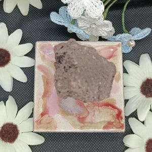 Peut inclure: Un carreau de céramique carré avec une finition marbrée rose, rouge et crème, surmonté d'une roche volcanique brun foncé, rugueuse et poreuse. Le carreau est entouré de plusieurs fleurs de marguerite artificielles blanches à cœur brun.