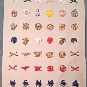 May include: A collection of vintage military insignia pins, arranged on a cream-colored paper. The pins feature various designs, including crossed rifles, eagles, anchors, and other emblems in colors like gold, silver, red, and blue. The pins are of varying sizes and shapes.