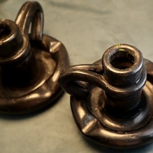May include: Two black ceramic candle holders with a ring handle. The holders have a circular base with a raised lip.