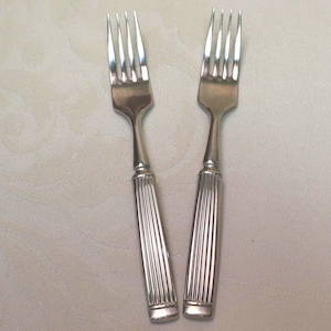 May include: Two silver forks with a classic design. Each fork has four tines and a handle with vertical grooves. The forks are displayed on a light-colored surface, showcasing their polished appearance.