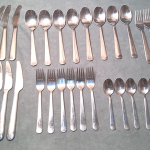 May include: A large set of stainless steel flatware including four dinner knives, four table knives, eight large spoons, six forks, and seven small spoons. Each piece features a smooth, polished silver finish with a simple, unadorned handle design.