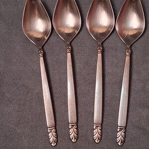May include: Four copper-toned metal spoons arranged in a vertical row. Each spoon features a smooth, elongated bowl and a straight handle that tapers slightly toward the base. The handle ends are decorated with a raised, symmetrical leaf-like pattern.