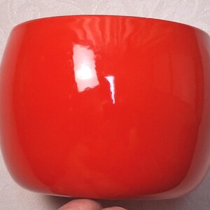 May include: A bright red, smooth, and glossy ceramic bowl with a rounded, bulbous shape. The surface of the bowl features a reflective finish that catches light, creating a distinct white highlight on its curved exterior wall.