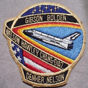 May include: An embroidered mission patch featuring a white space shuttle against a light blue background with a rainbow arc. The patch includes the names GIBSON, BOLDEN, NELSON, HAWLEY, CHANG-DIAZ, and CENKER NELSON, along with stars and a US flag motif.